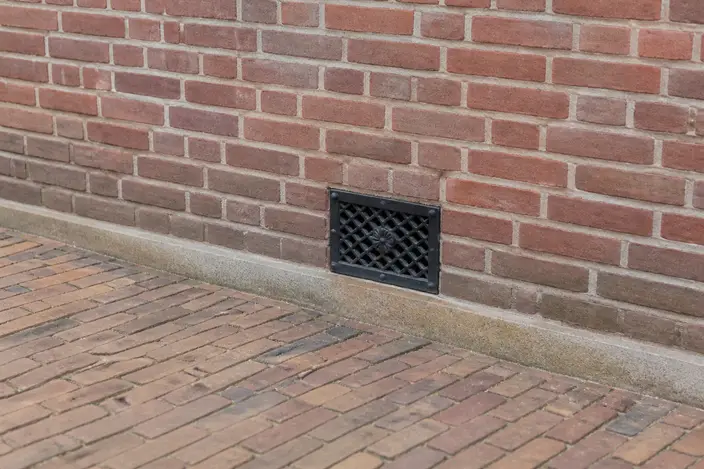 AI-beeld: de onderkant van een oude rode bakstenen muur boven een bestrate stoep, met een zwart gietijzeren ventilatierooster van de kruipruimte