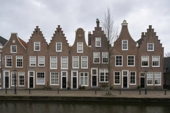 AI-beeld: een rij oude bakstenen huizen met trapgevels en halsgevels en witte kozijnen aan een gracht, op een bewolkte ochtend